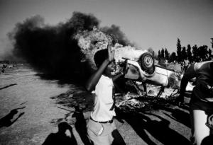 As a car burns behind him, a young South African participates in a civil disturbance outside the Aud